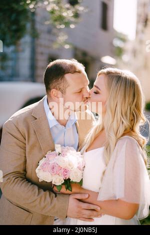 The bride with bouquet and groom are embracing and kissing on a cozy street of the old city, close-up Stock Photo