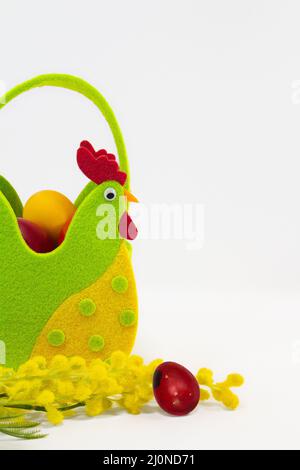 Felt rooster basket with painted eggs and mimosa flowers on white background. Easter. Copy space Stock Photo