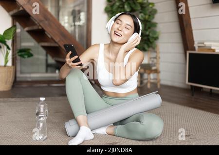 Happy asian girl in fitness clothing sitting at home on floor, listening music and workout, holding smartphone and singing durin Stock Photo