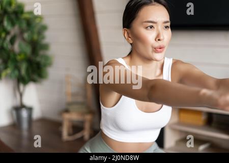 Young asian fitness woman with healthy fit body, doing squats, morning workout, wearing activewear, standing at home in living r Stock Photo