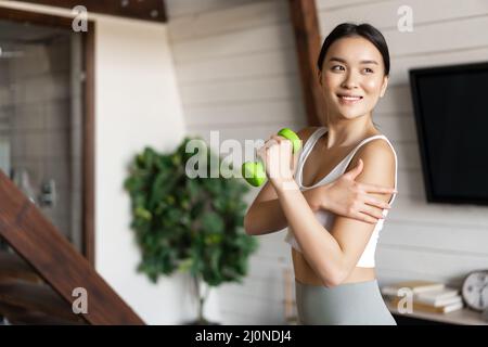 Active and healthy asian girl with fit body doing fitness exercises at home, lifting dumbbells and touching her biceps, workout Stock Photo