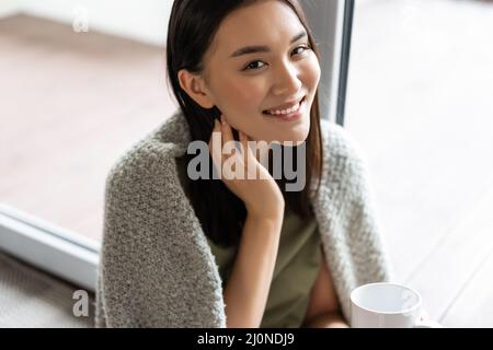 Portrait of beautiful asian woman wrapped in blanket, drinking coffee and smiling, relaxing at home on weekend, sitting near win Stock Photo