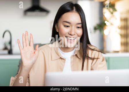Smiling asian girl student waving hand at laptop camera. Woman on video call from home, saying hello on videochat, sitting in ki