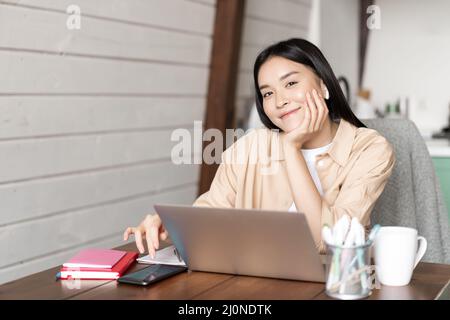 Smiling asian woman working from home remotely, sitting with laptop and mobile phone. Korean girl doing homework, e-learning wit Stock Photo