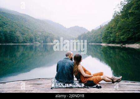 Man with pregnant woman are sitting on a pier by the lake in the park Stock Photo