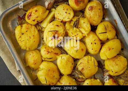 Oven baked whole crushed and crusty potato spuds with seasoning and herbs in metalic tray Stock Photo