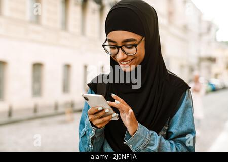 Young muslim woman wearing hijab smiling and using mobile phone at city street Stock Photo