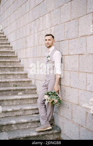 Groom with a bouquet stands on the stone steps, leaning his back against the wall Stock Photo