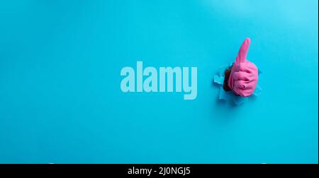 Hand in a pink household rubber glove sticks out of the torn hole and shows a gesture okay, thumb up. Blue background Stock Photo