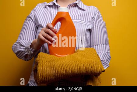 A woman in a white striped shirt holds an orange plastic bottle with liquid washing gel and a knitted sweater. Laundry and house Stock Photo