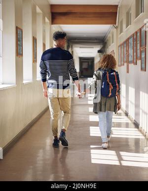 He walks her to every class. Rearview shot of an affectionate young couple walking hand in hand through their university corridor. Stock Photo