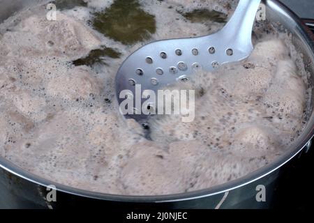 Meat broth with abundant foam boils in saucepan, foam is removed with metal slotted spoon. Cooking soup on domestic kitchen. Selective focus. Close-up Stock Photo