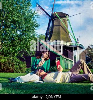 Vintage Netherlands 1970s, couple of middle aged tourists relaxing in meadow beside a windmill, Holland, Europe, Stock Photo