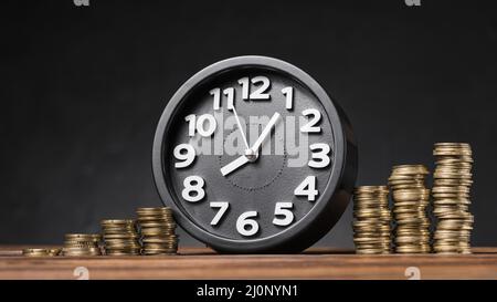 Round clock between the increasing coins on wooden desk against black backdrop . High quality and resolution beautiful photo con Stock Photo