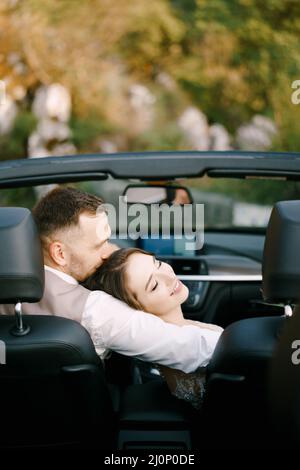 Groom hugs bride who has laid her head on his shoulder in a convertible Stock Photo