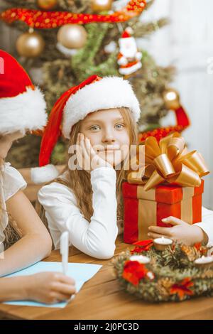 Little Sisters Girls in Christmas Hats Write a Letter to Santa Claus and Enjoy Christmas Eve. Stock Photo