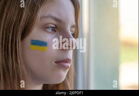 War. Russia's invasion of Ukraine. Portrait of a child with tears in his eyes sitting on the table and looking out the window. The blue and yellow Stock Photo
