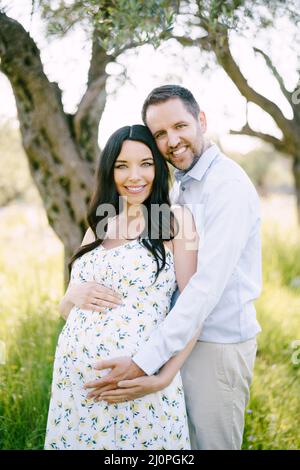 Smiling man hugging pregnant woman in green park Stock Photo