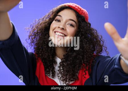 Cool and stylish friendly-looking energized happy woman in 20s with curly hair wearing warm beanie and outdoor clothes tilting h Stock Photo