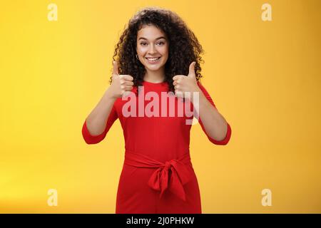 Excellent idea, nice job. Portrait of supportive delighted and happy charming female friend showing thumbs up as standing in red Stock Photo
