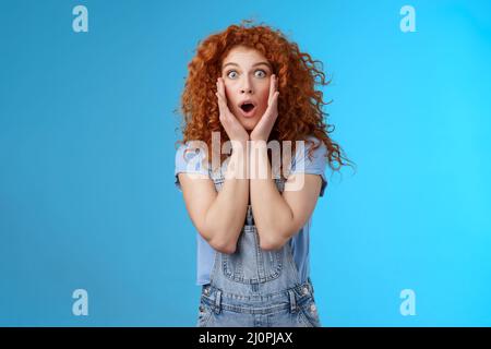 Amazed gasping shocked wondered redhead attractive curly girl popping eyes stare camera drop jaw concerned astonished touch chee Stock Photo