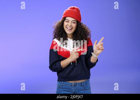 Charismatic playful young curly-haired european female winking joyfully at camera as pointing at upper right corner wearing hat Stock Photo