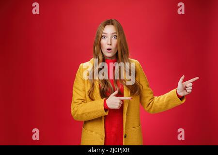 Speechless questioned and confused surprised redhead girl in yellow autumn coat pointing right open mouth curiously and staring Stock Photo