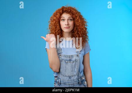 Silly young happy cute redhead woman curly hairstyle smirking rate fine place hang out pointing left thumb introduce not bad cho Stock Photo