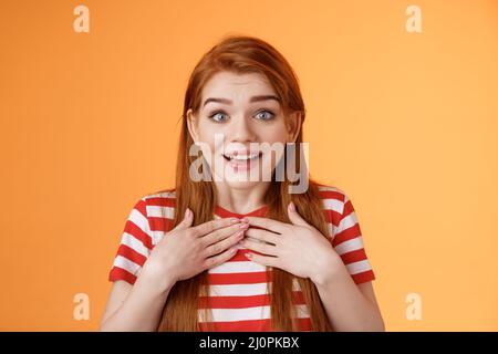 Redhead cute girl receive pleasant news. Charmed happy ginger woman press hands chest astonished surprised, look camera delighte Stock Photo