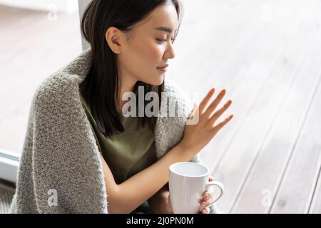 Thoughtful asian woman sitting at home on floor, touching glass window nostalgic, yearning to go outside, being on self-quaranti Stock Photo