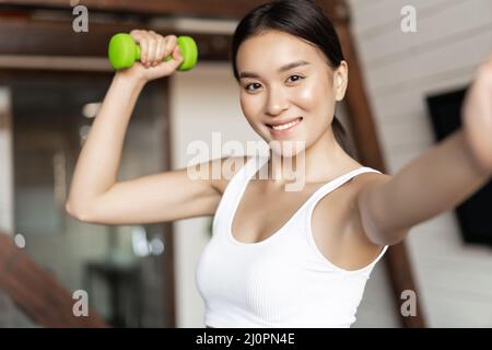 Smiling korean girl taking selfie with dumbbell, workout at home during pandemic, wearing activewear Stock Photo