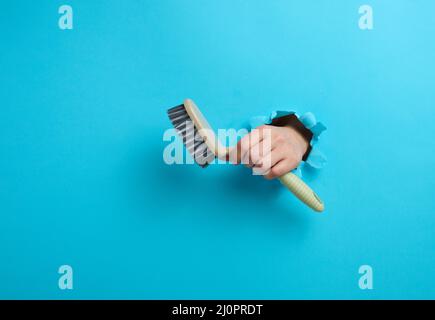 Female hand holds a gray plastic brush on a blue background. A part of the body sticks out of a hole with torn edges in a paper Stock Photo