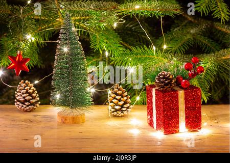 A red Christmas box with a gift, a red star and a small Christmas tree on the background of a natural fir tree with Christmas li Stock Photo