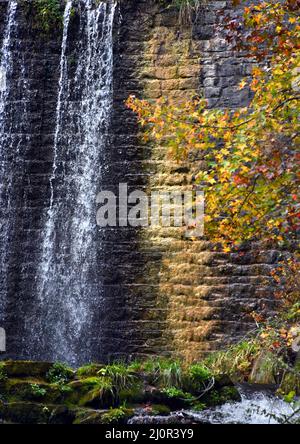 Mirror Lake Dam has colorful stone wall.  Yellow and orange Fall leaves add to the color.  Water is spilling over dam. Stock Photo