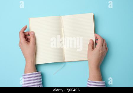 Two female hands hold an open notebook with blank white sheets on a blue background, top view Stock Photo