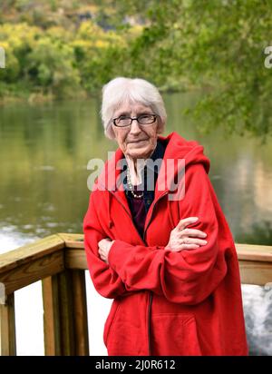 Elderly woman stands on wooden dock overlooking the White River in North Arkansas.  She is wearing a red hoodie and has white hair. Stock Photo
