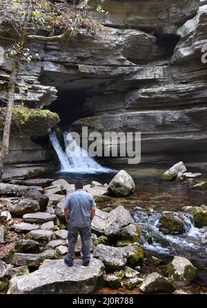 Visitor, to Blanchard Springs Caverns, stands on a large rock admiring the waterfall.  He has on grey pants and shirt and has hands in his pockets. Stock Photo