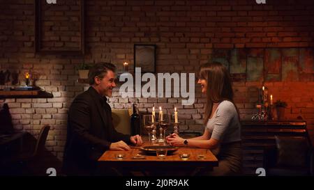 Elegant couple having romantic dinner at home, sitting at table in living room drinking red wine, talking. Stay at home concept. Stock Photo