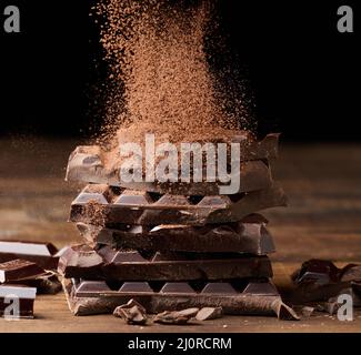 Cocoa powder sprinkles on a stack of broken dark chocolate on a wooden table, black background Stock Photo