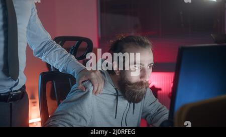 Bearded long-haired hippie looking man playing games at the computer, employer man puts his hand on hippie looking teenager's shoulder asking what he Stock Photo