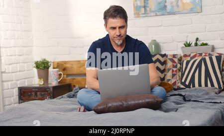 Casual man sitting on bed working online from home with laptop computer. Stay at home and work in home office. Stock Photo