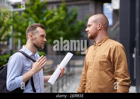 Male caucasian architects or designers holding drawings or blueprints making conversation about project, brainstorming. Business people talking outdoor modern building background. High quality image Stock Photo