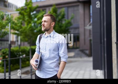 Cute bearded caucasian businessman, real estate agent, designer or engineer standing outdoor with laptop and plans. Modern building and trees on background. High quality image Stock Photo