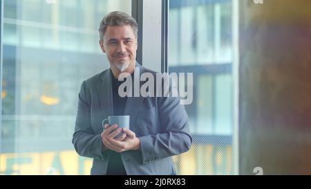 Happy older businessman taking break in office drinking coffee standing in window and daydreaming, thinking. Stock Photo