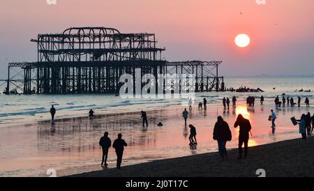 Brighton UK 20th March 2022 - Crowds watch the sun setting behind Brighton's West Pier on the Spring Equinox as warm sunny weather is forecast to continue throughout Britain over the next week : Credit Simon Dack / Alamy Live News Stock Photo