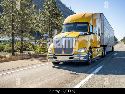Classic long haul big rig yellow semi truck tractor with truck driver cab sleeping compartment transporting cargo in refrigerator semi trailer running Stock Photo