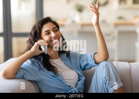 Cheery young brunette lady sitting on sofa, having mobile phone conversation at home, copy space Stock Photo