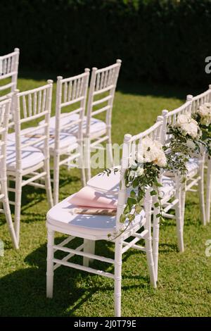 Row of white chairs decorated with bouquets of flowers stands on a green lawn Stock Photo