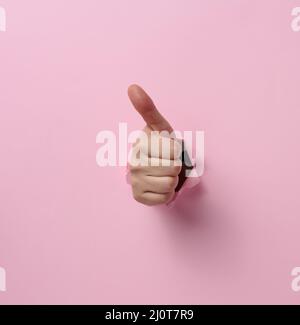 A woman's hand sticks out of the torn hole and shows a gesture okay, thumb up. Pink background Stock Photo