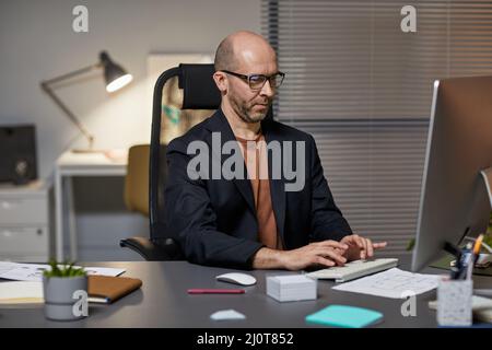 Portrait of bald adult businessman using computer at workplace while working late at night in office Stock Photo